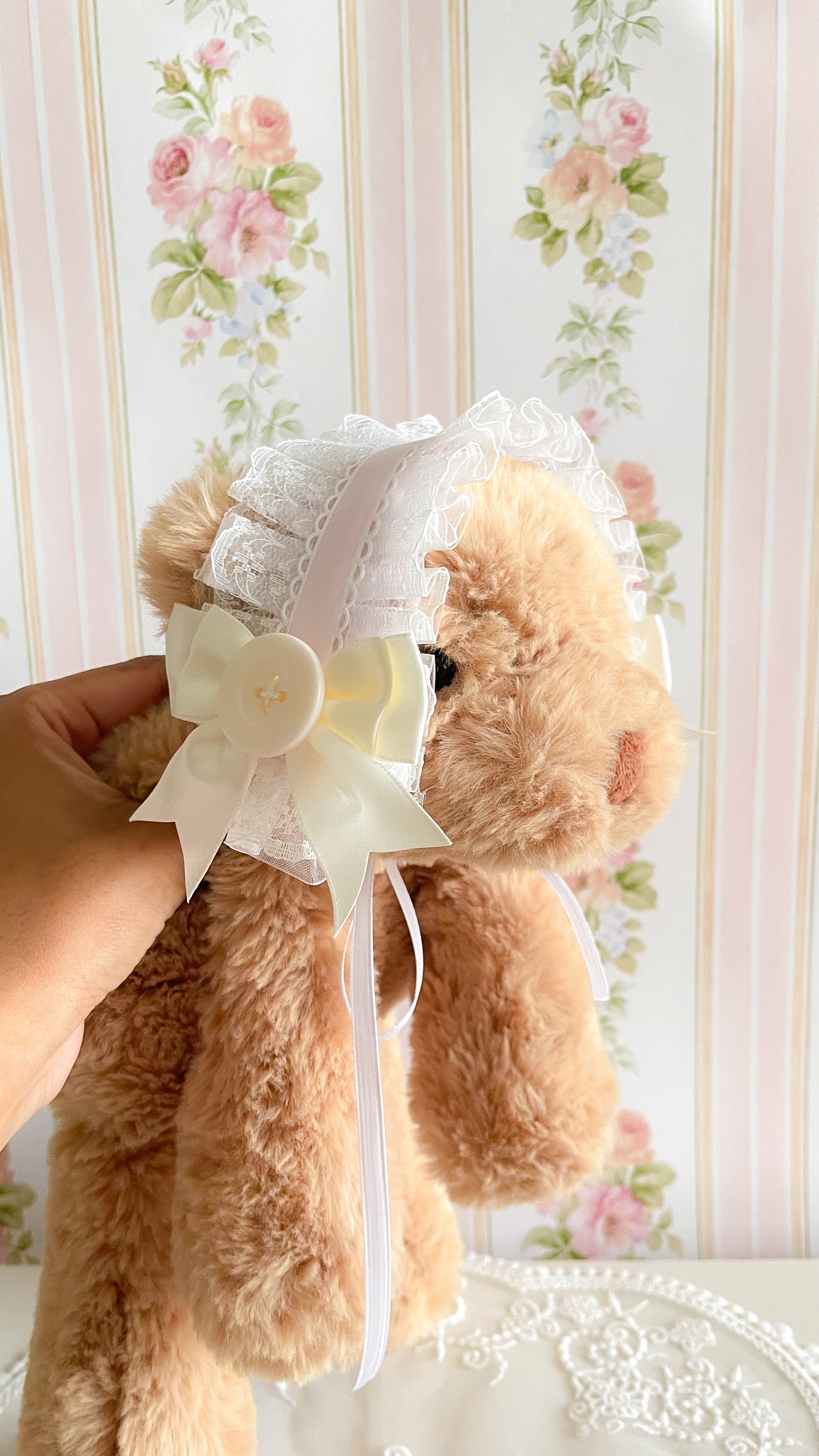 Teddy bear with a white bow and decorative headband against a floral wallpaper background